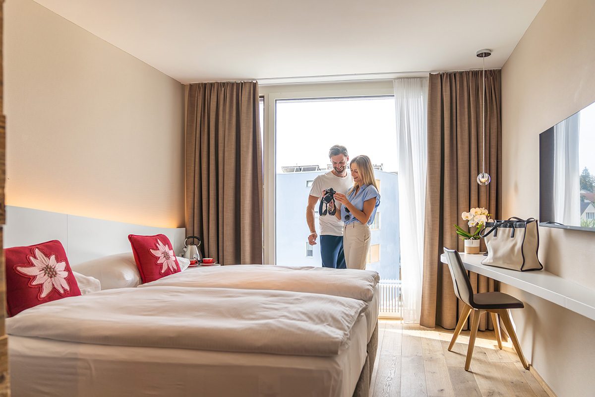 Lodge Zimmer mit Paar, Hotel Allegra Lodge, Doppelbett mit roten Kissen, dahinter Paar am Fotos auf der Kamera ansehen, rechts Holzkonsole mit Stuhl und darüber TV-Gerät