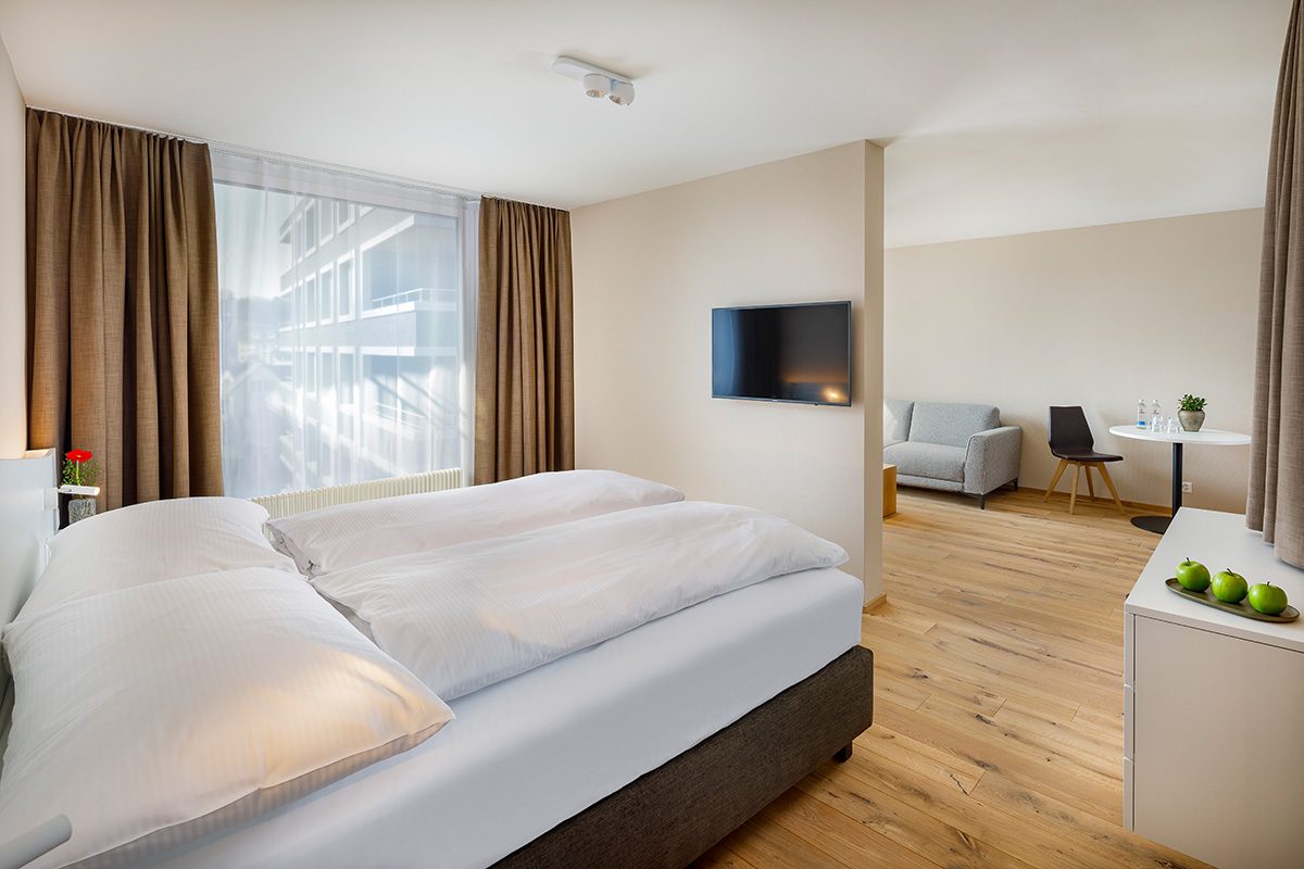 achesa homes, Serviced Apartments, Doppelbett mit weisser Bettwäsche, daneben Kommode mit Äpfeln und davor TV-Gerät an der Wand, im Wohnraum Sofa mit Tisch