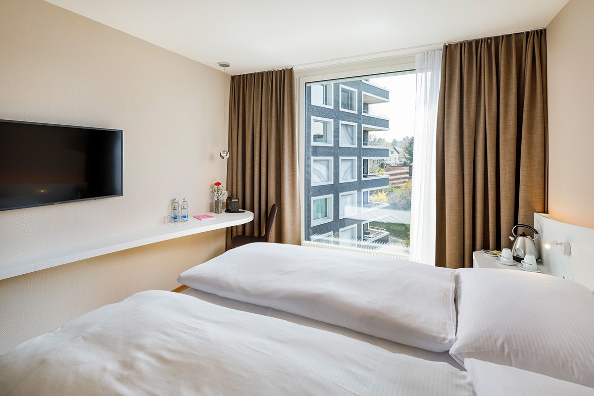 Tageszimmer, Hotel Allegra Lodge, Doppelbett mit weisser Bettwäsche und roten Kissen mit weissem Edelweiss, auf dem Nachttisch stehen Wasserflaschen und ein Thermoskrug mit Tassen, vor dem Bett gibt es ein weisses Ablageboard und darüber ein TV-Gerät
