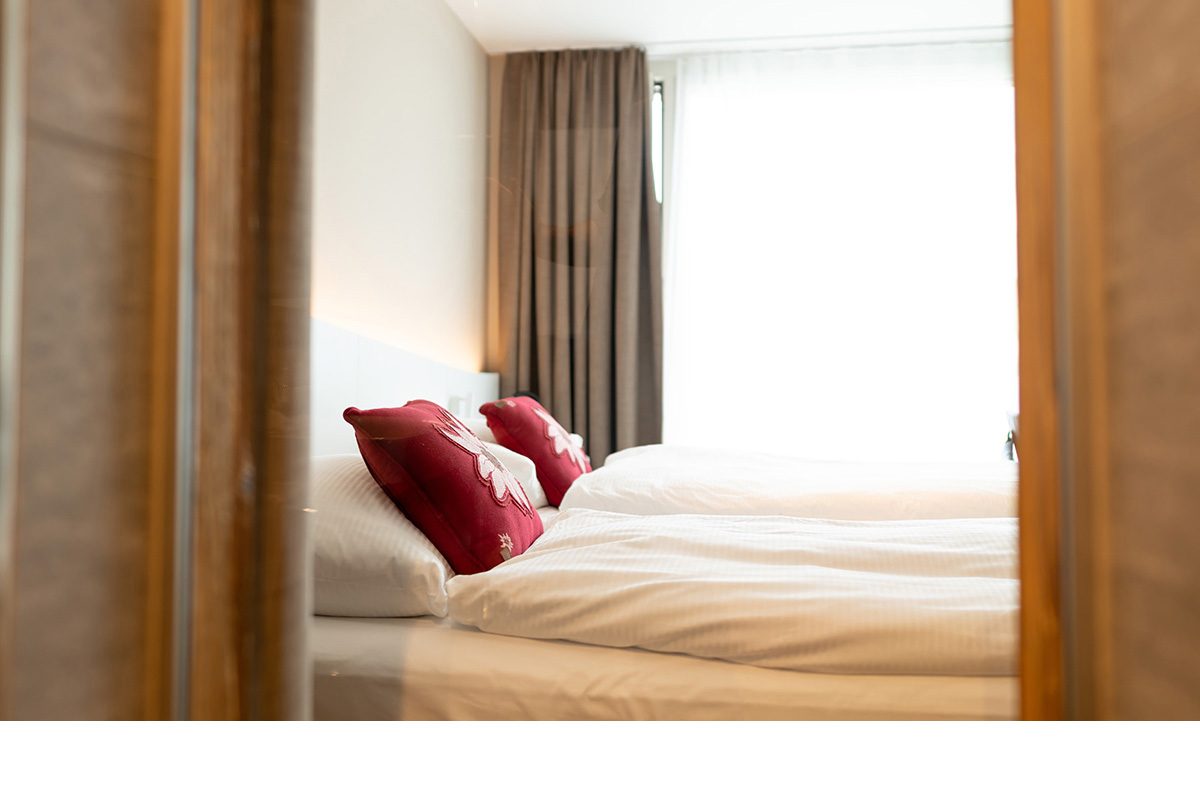 Blick durch einen Türrahmen auf ein helles, modern eingerichtetes Hotelzimmer mit Doppelbett, weissn Kissen und dekorativen roten Zierkissen mit Edelweiss.