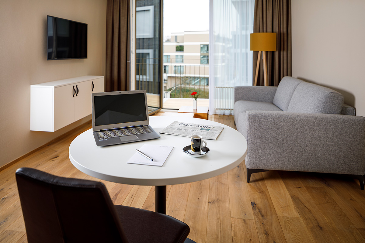 Grosses Wohnzimmer mit Arbeitsplatz und im Hintergrund wird der Balkon mit offener Tuer gezeigt.