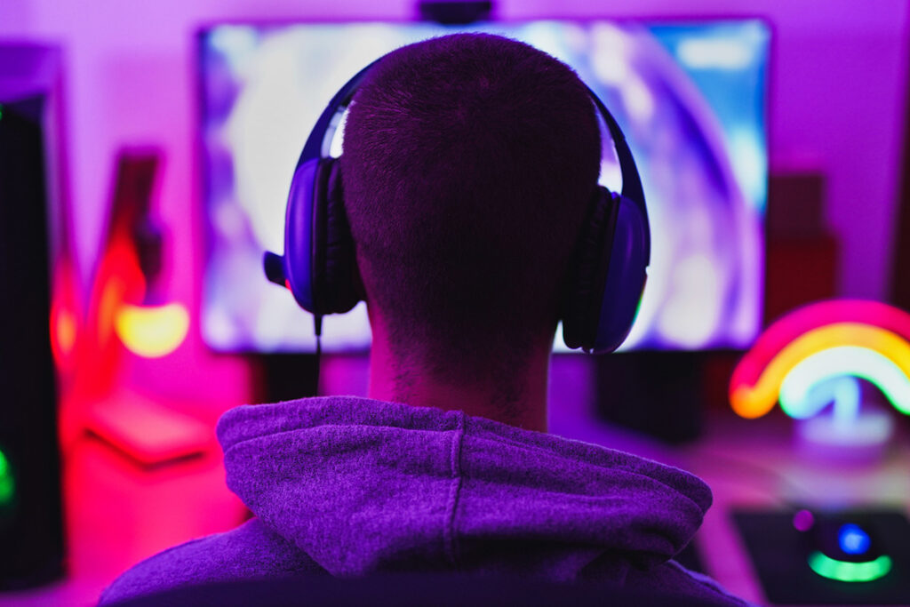 Rückansicht eines Gamers mit Headset in einem stimmungsvoll in Lila und Pink beleuchteten Raum vor einem Monitor – passend zum Gamer-Package der Allegra Lodge