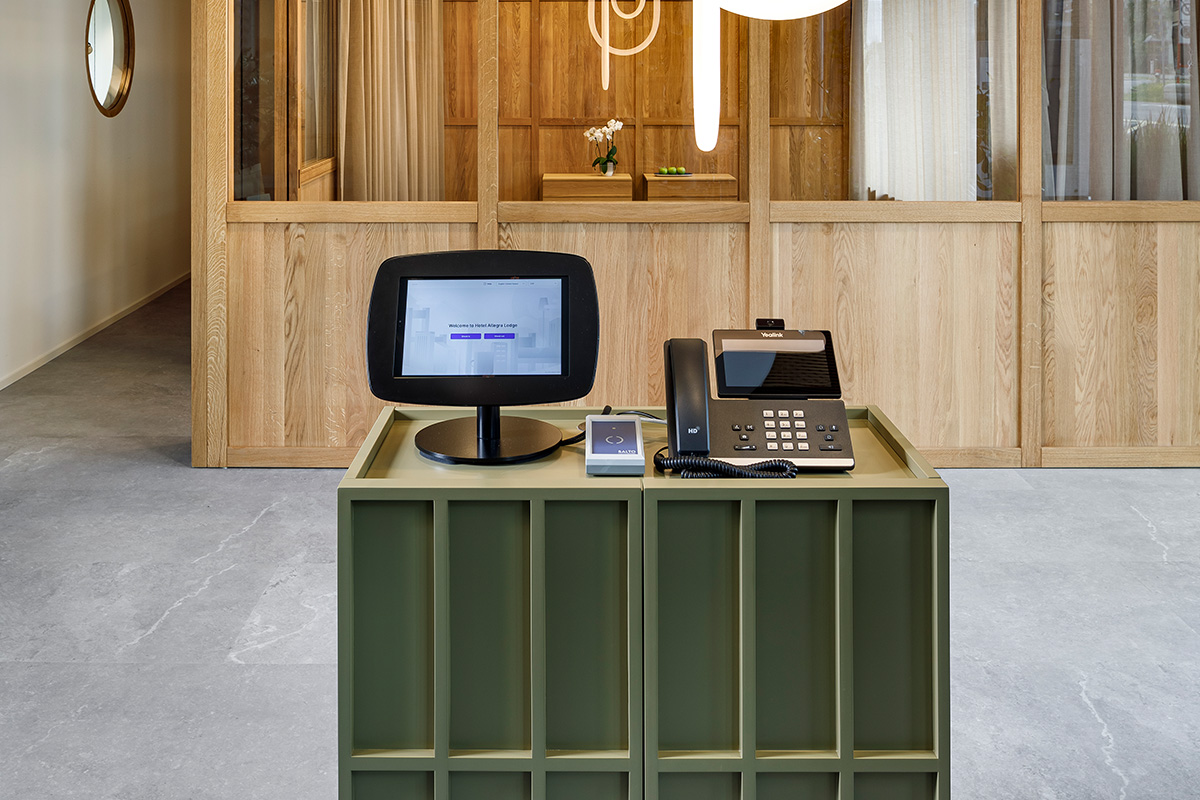 Moderner Self-Check-in-Terminal der achesa Group im Hotel Allegra Lodge mit Touchscreen-Monitor, Kartenlesegerät und VoIP-Telefon auf einem olivgrünen Standfuss.