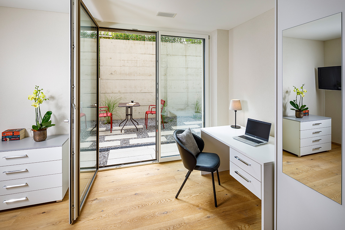 Ein heller Arbeitsplatz in einem modernen Co-Living Zimmer mit Eichenparkett. Ein weisser Schreibtisch mit Laptop und schwarzem Polsterstuhl steht neben einer grossen, weit geöffneten Glastür. Der Blick führt direkt auf eine private Terrasse mit hellen Steinplatten auf Kies, zwei roten Metallstühlen und einem kleinen Tisch vor einer Sichtbetonwand.
