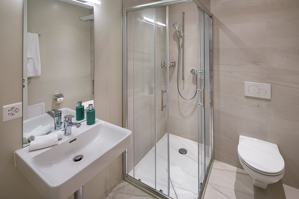 Ein helles, modernes Badezimmer mit beigen Fliesen. Zu sehen ist eine Glas-Duschkabine mit verchromten Armaturen, ein weisses Waschbecken mit grossem Spiegel und zwei grünen Seifenspendern sowie ein wandhängendes WC. Das Design ist schlicht und funktional, ergänzt durch frische weiße Handtücher.