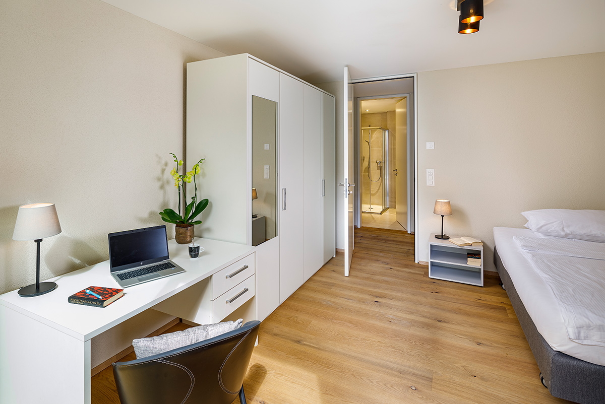 Co-Living Zimmer im Allegra Lodge Style in Kloten: Helles Zimmer mit Arbeitsbereich, modernem Bad (im Spiegel sichtbar) und direktem Zugang zum privaten Gartensitzplatz mit modernen roten Sitzmöbeln.